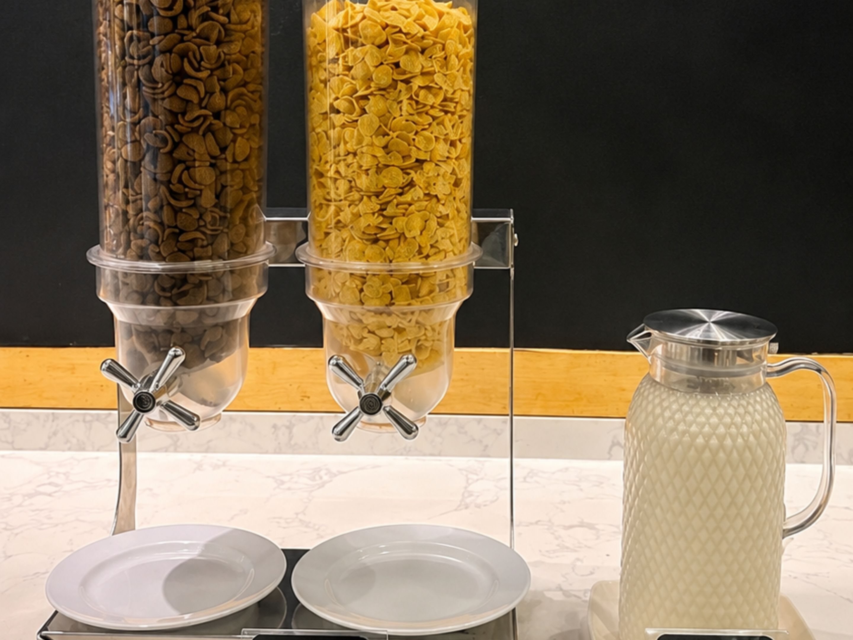 A buffet setup with two dispensers, one for cereal and one for a beverage, and a pitcher on the side.