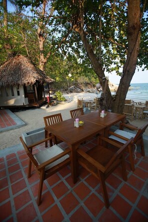A wooden table with chairs set on a tiled patio, overlooking a beach with a thatched-roof structure in the background. - Thongtapan Resort (Ko Pha-ngan)
