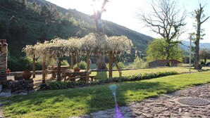 Un entorno rústico al aire libre con un muro de piedra, una pérgola de madera y una zona verde bajo un cielo despejado.