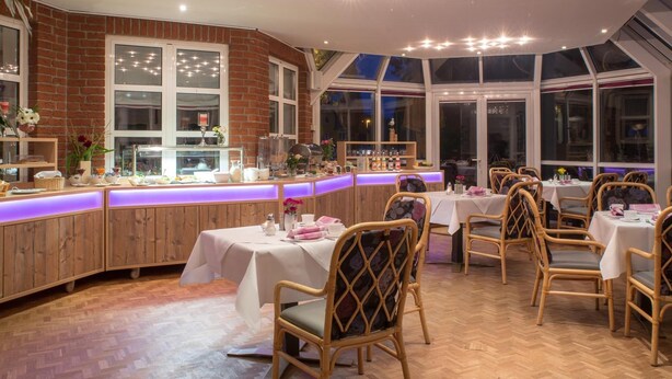 Un restaurante con paneles de madera, una zona de bar con mostrador iluminado en morado y mesas con manteles blancos y sillas de mimbre.