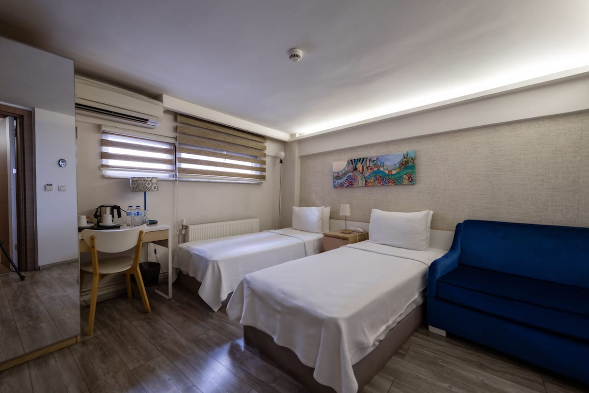 Habitación de hotel con dos camas individuales, un escritorio con una silla, un sofá azul y un cuadro en la pared.