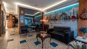 Un hall de hotel con suelo de baldosas, un sofá de cuero negro, una mesa de centro de madera y un mural con paisajes urbanos en la pared.