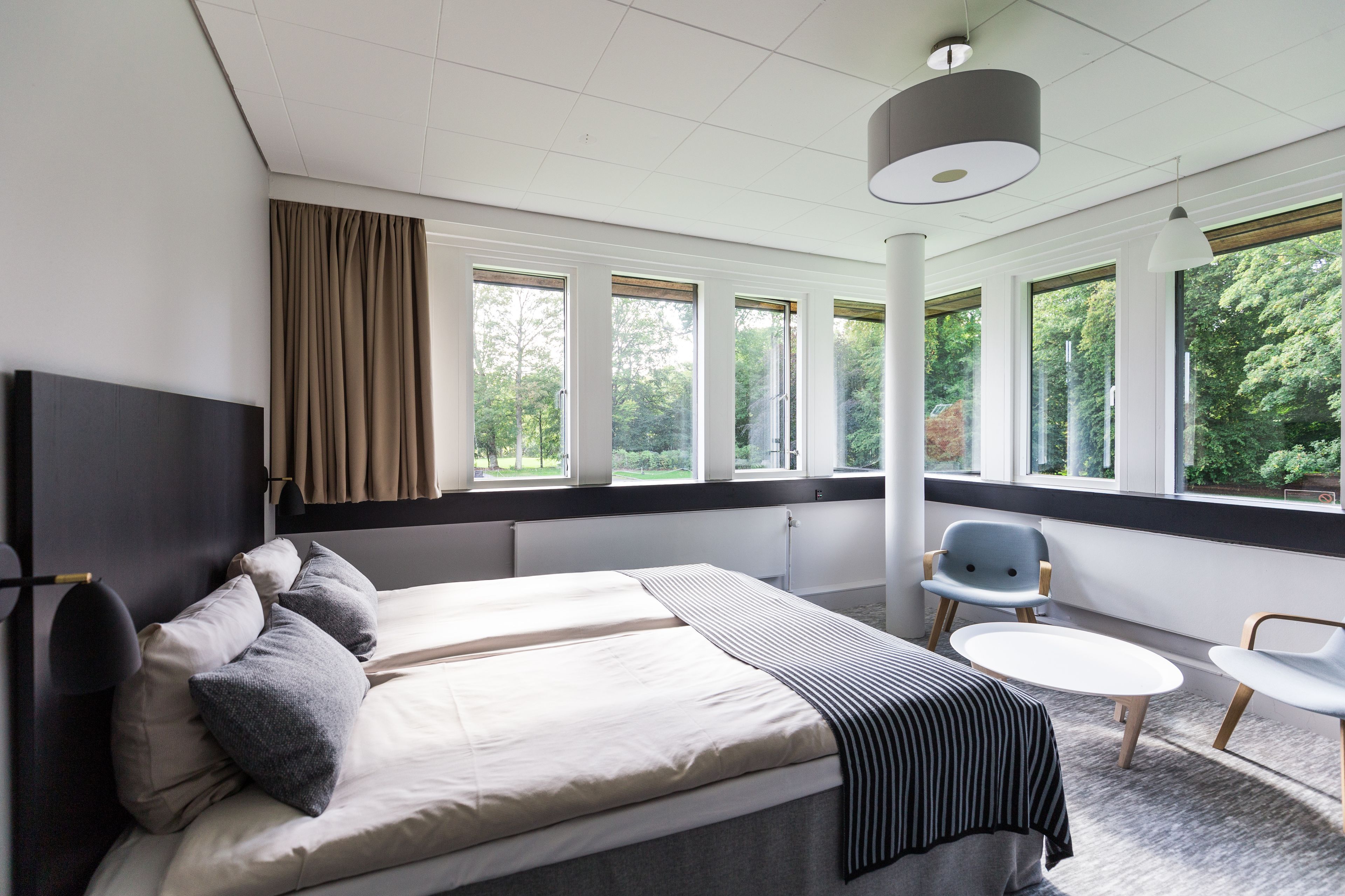 A modern hotel room with a large bed, a small table, and a chair. There are multiple windows providing a view of the outdoors.