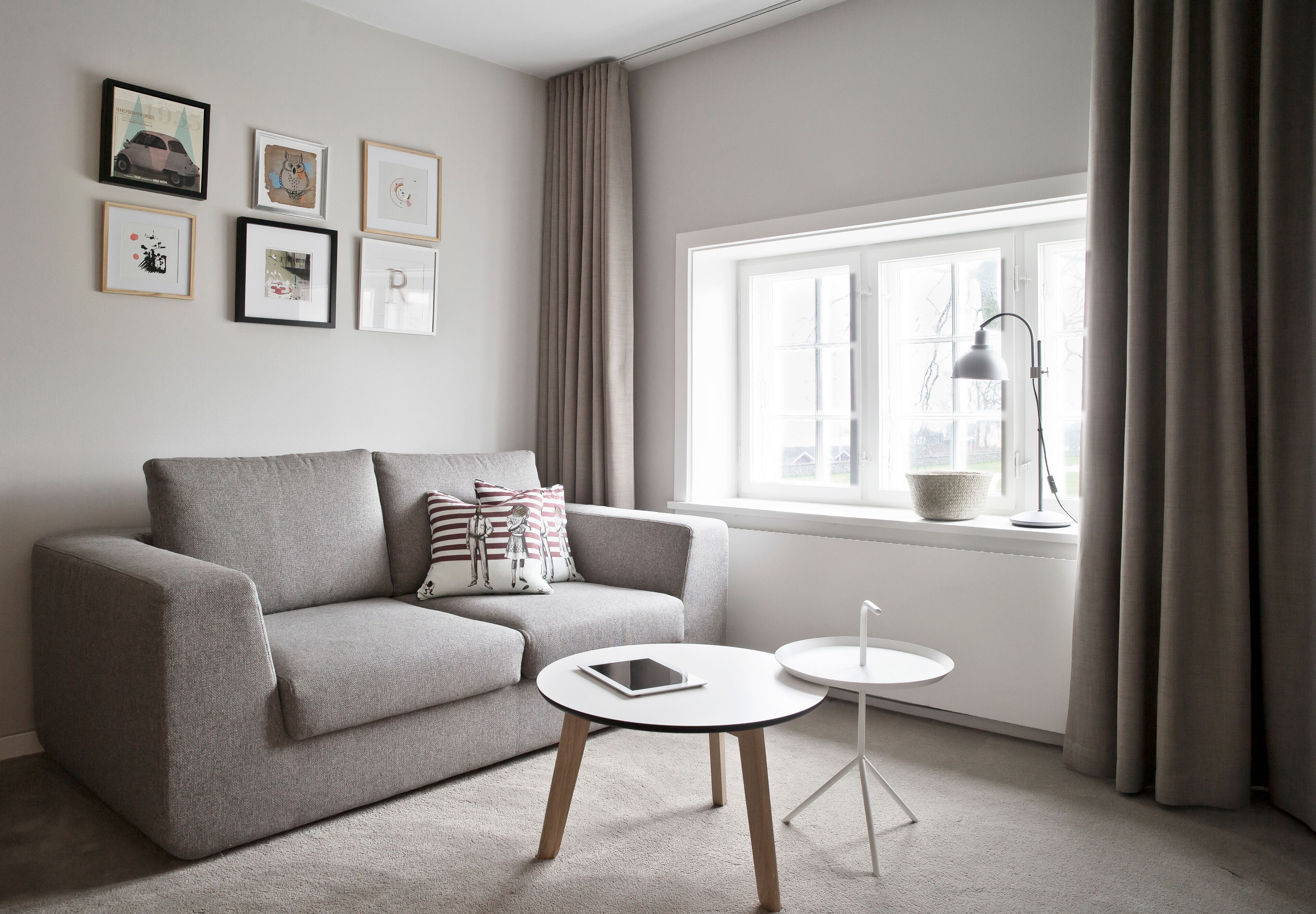 A modern living room with a grey sofa, a round coffee table, and a side table. There are framed pictures on the wall and a window with curtains.