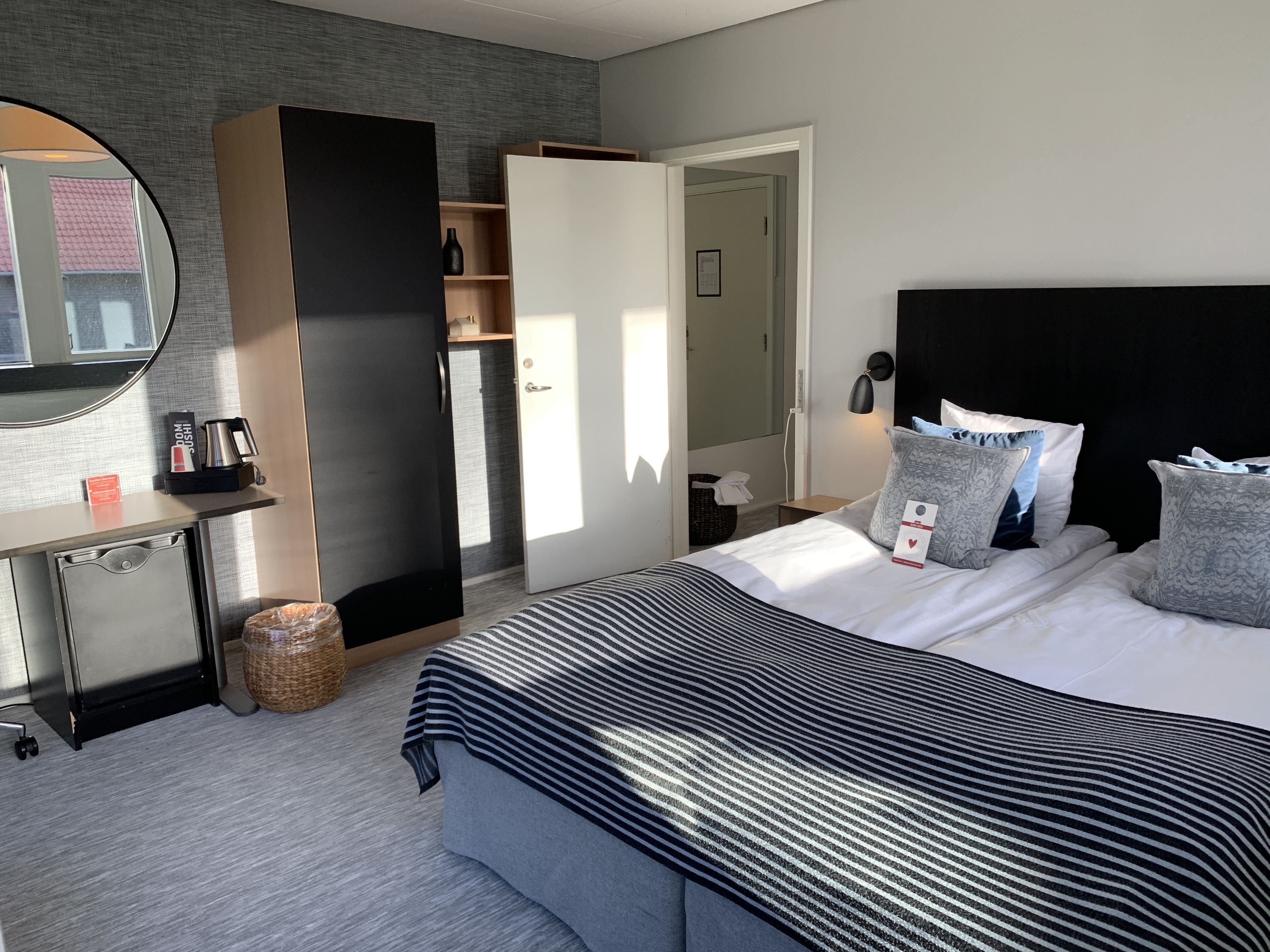 A modern hotel room with a large bed, a wardrobe, a small desk, and a mirror.