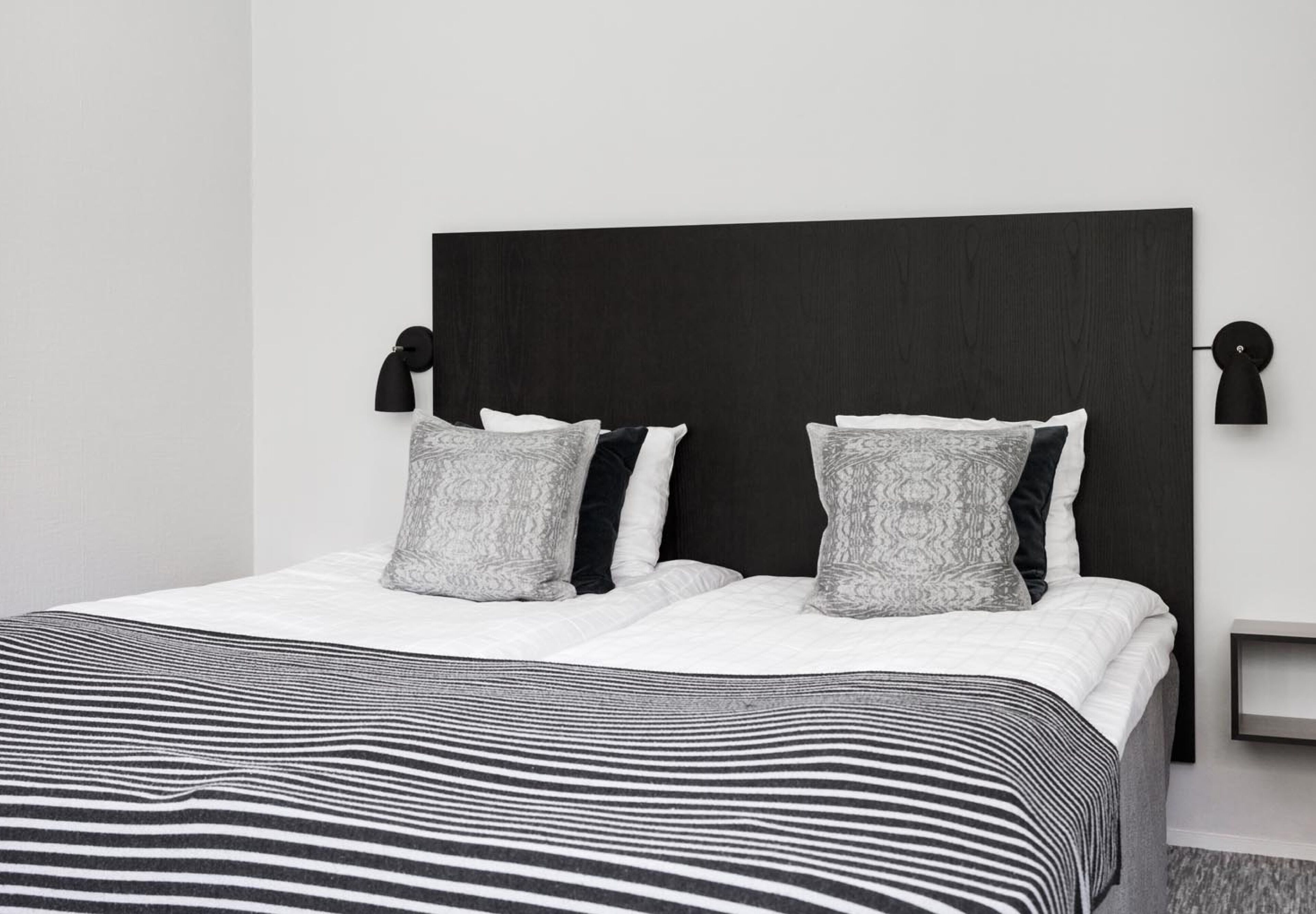 Une chambre d’hôtel moderne avec un grand lit, une tête de lit sombre et deux lampes fixées au mur.