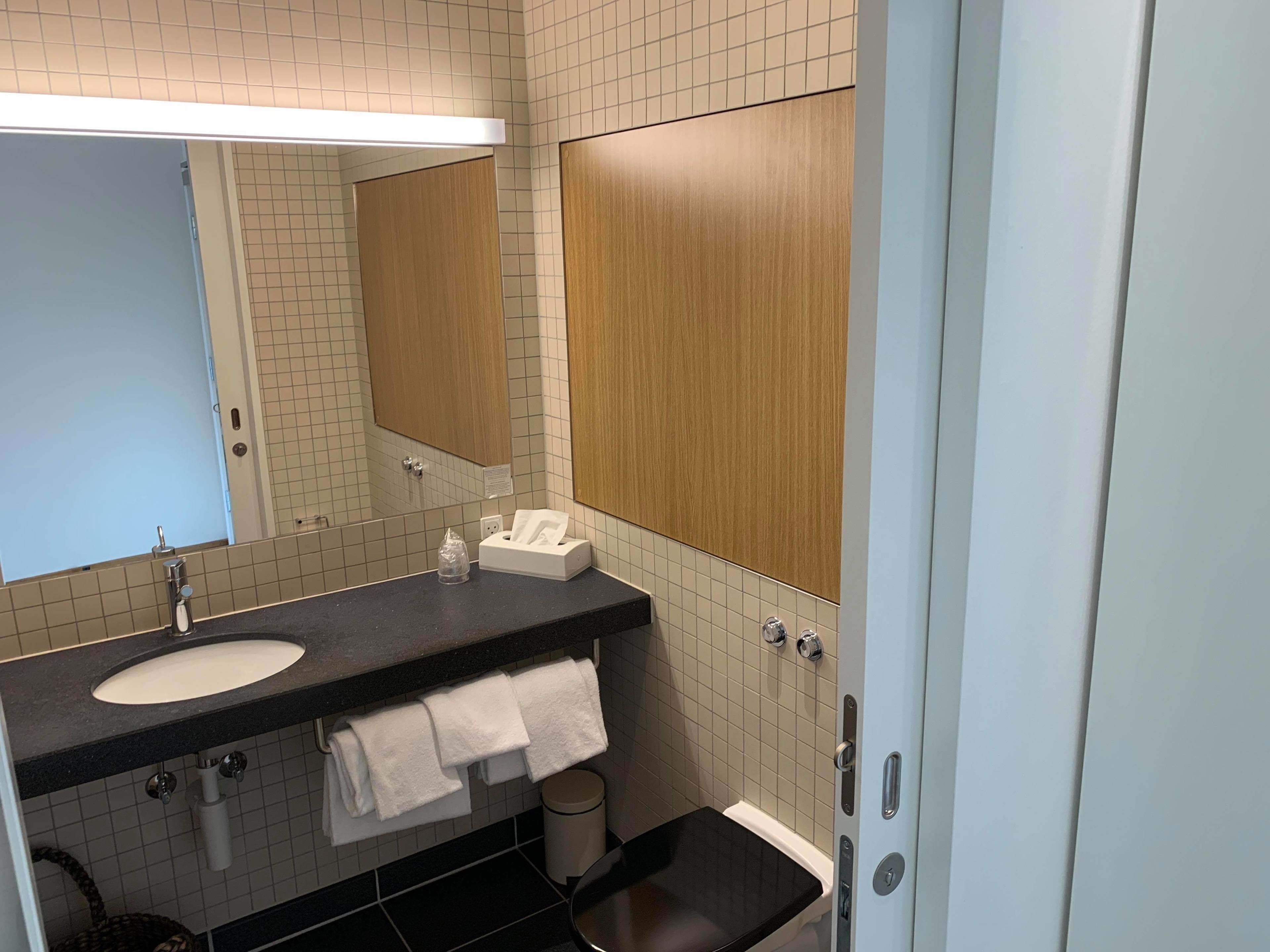 Un baño moderno con lavamanos, espejo, dispensador de toallas y un cubo de basura.
