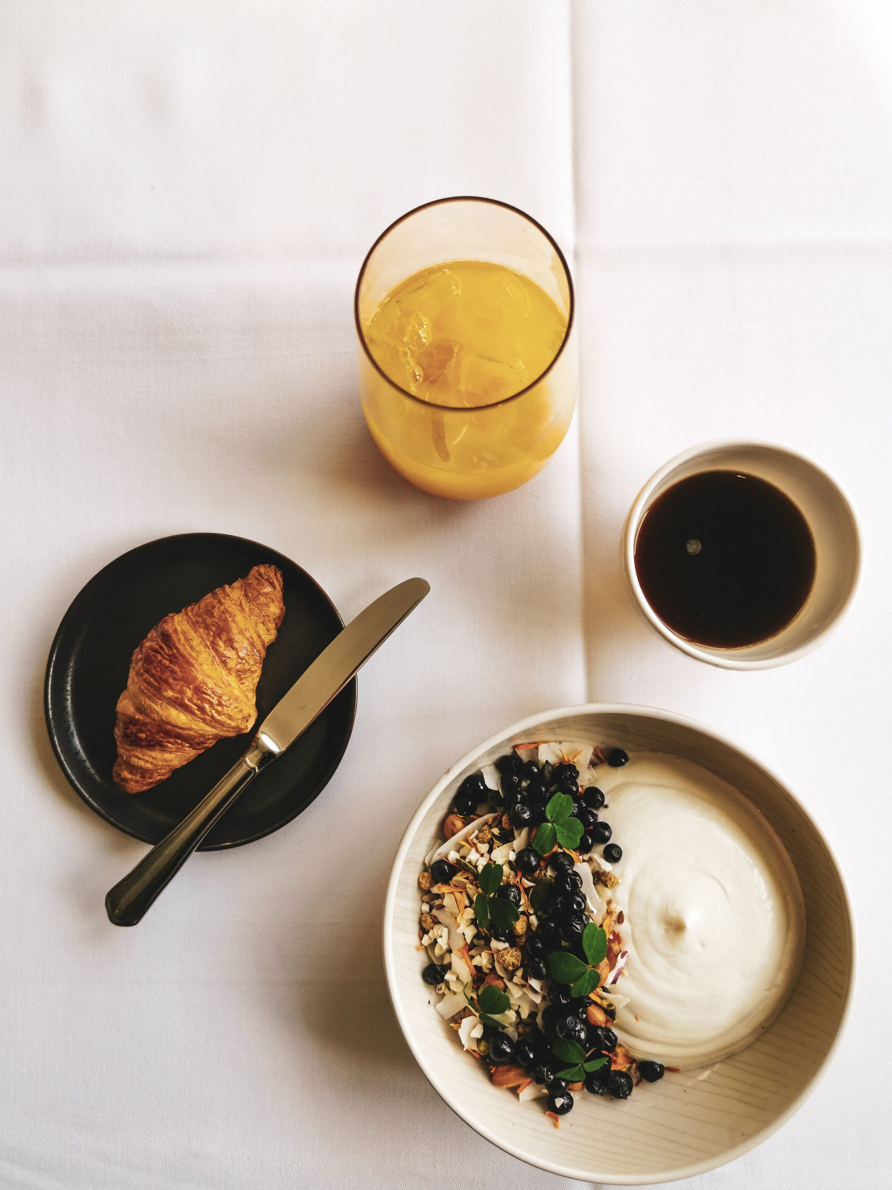 A breakfast setting with a croissant, a glass of orange juice, a bowl of yogurt with berries and nuts, and a cup of coffee on a white tablecloth.