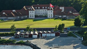 A large, historic building with a prominent flag, surrounded by well-maintained lawns and a sandy area with sports equipment. - Rungstedgaard (Rungsted Kyst)