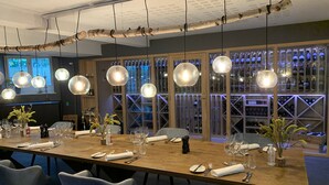 A long dining table set for a meal with multiple chairs, hanging pendant lights, and a wine rack in the background. - Rungstedgaard (Rungsted Kyst)