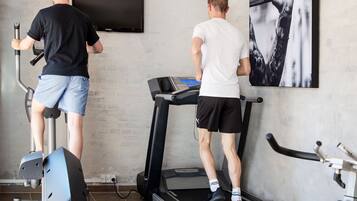 Two people exercising on stationary bikes and treadmills in a gym with a large wall-mounted TV and a framed picture.