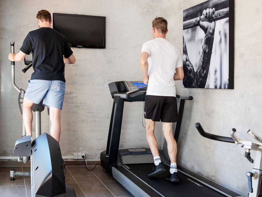 Two people exercising on stationary bikes and treadmills in a gym with a large wall-mounted TV and a framed picture.