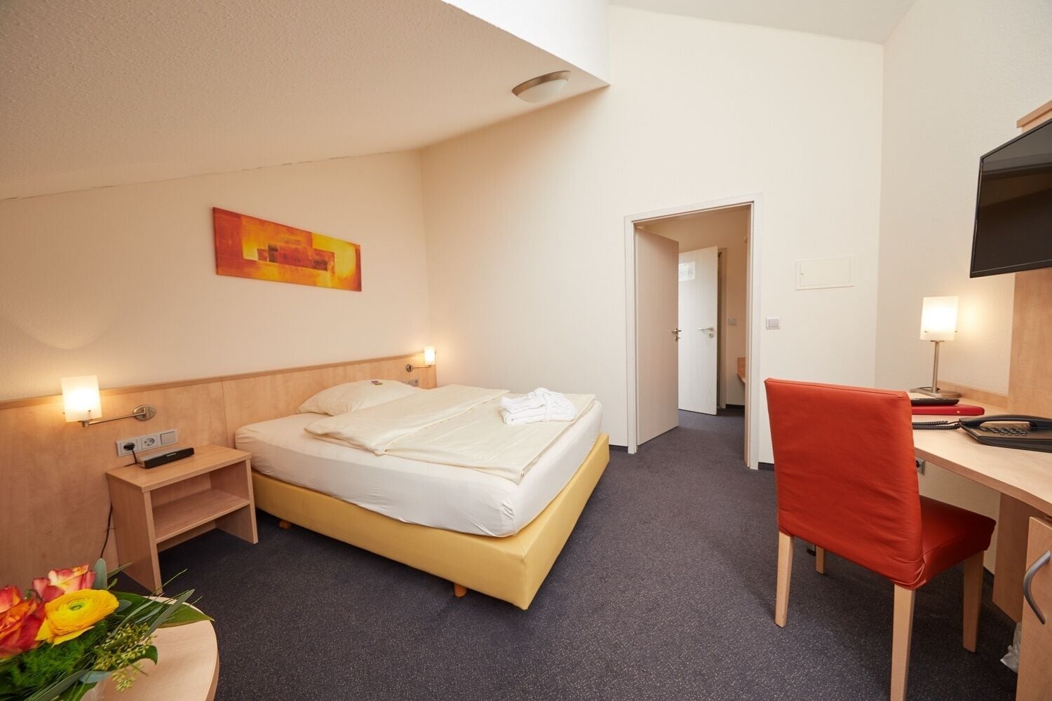 Ett hotellrum med en säng, ett sängbord, en lampa, ett skrivbord, en stol och en tv.