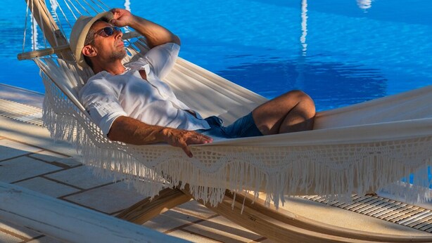 Un hombre descansando en una hamaca junto a una piscina con sillones de playa y sombrillas al fondo.