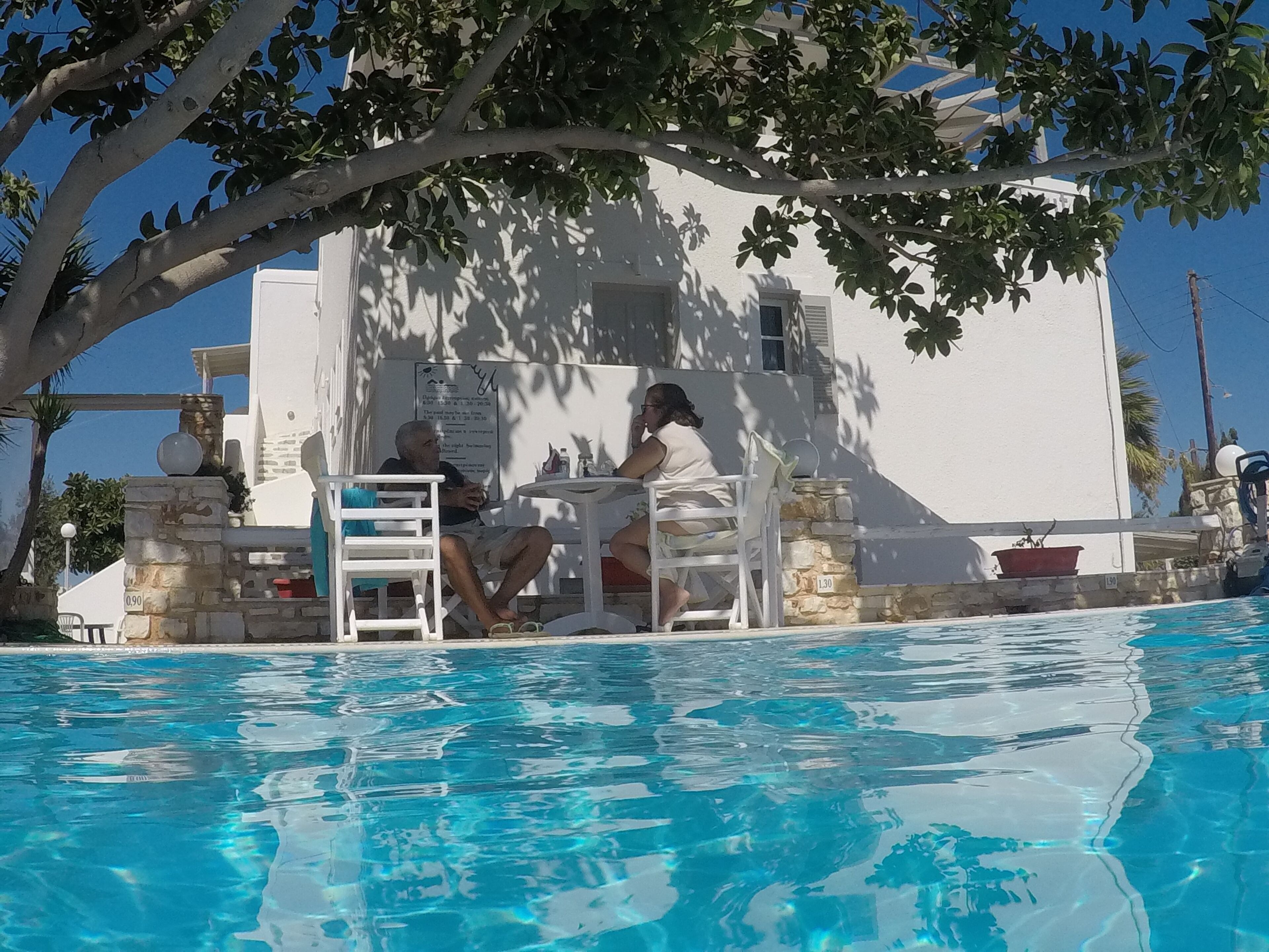 A poolside area with two people seated at a table under a tree, a white building in the background, and a clear blue pool.