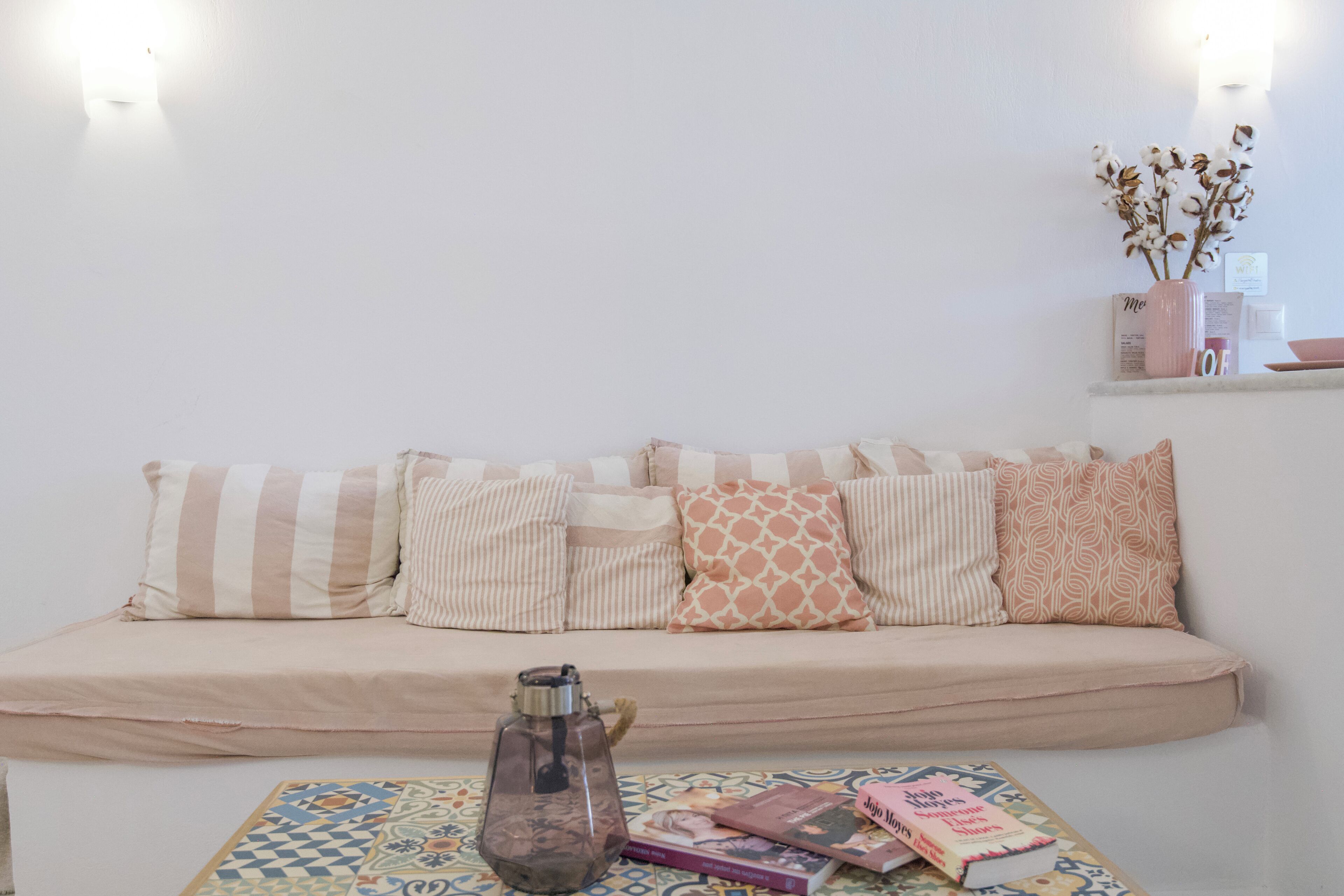 A cozy room with a sofa adorned with striped and patterned cushions, a small table with a decorative vase and books, and a wall-mounted shelf with a vase of flowers.