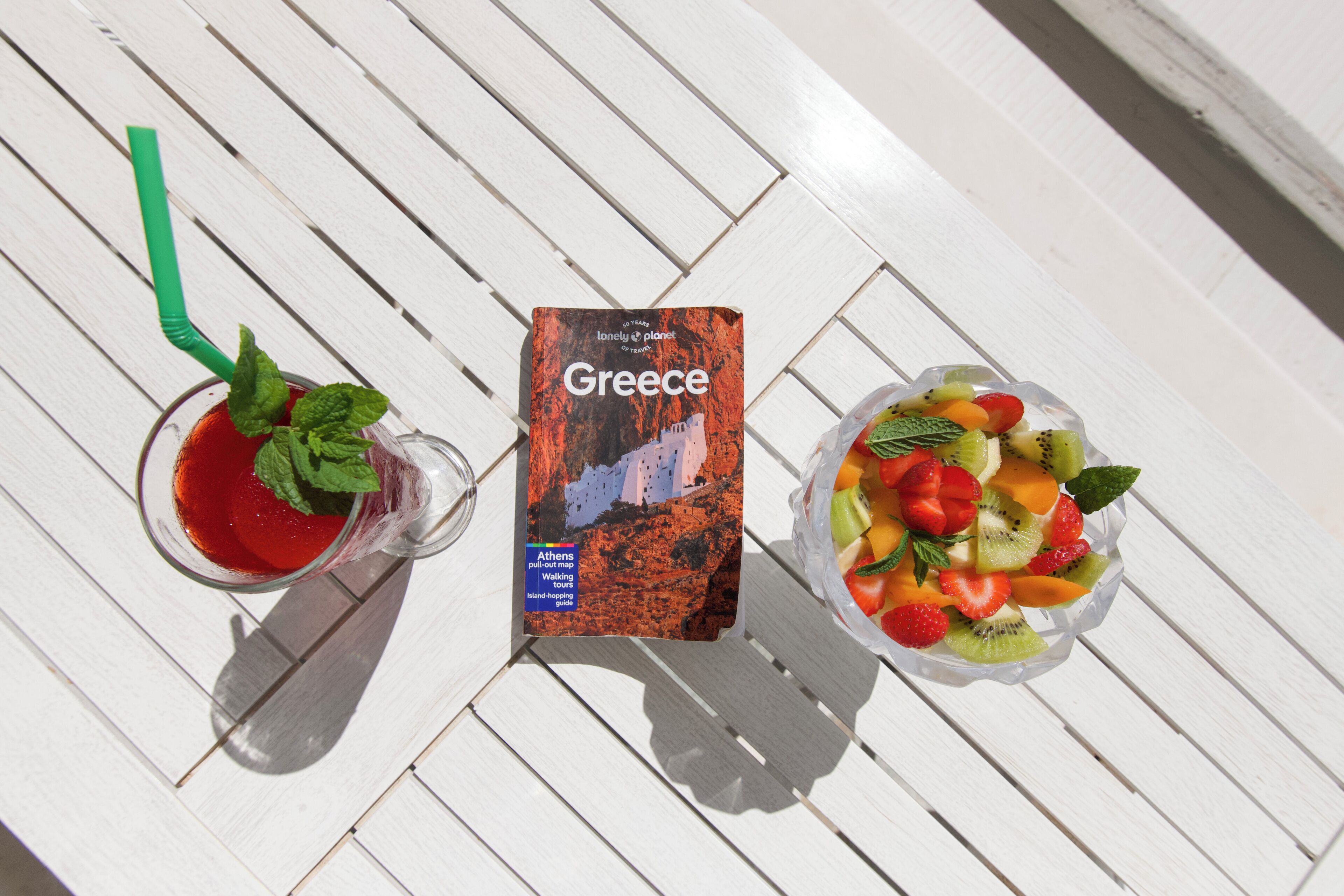Eine herausziehbare Karte von Athen, Griechenland, ein Glas rotes Getränk mit Minze und eine Schüssel mit gemischtem Obst auf einem weißen Holztisch.