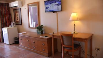 Een hotelkamer met een houten bureau, een stoel, een aan de muur bevestigde televisie, een lamp, een dressoir, een potplant en een raam met gordijnen.