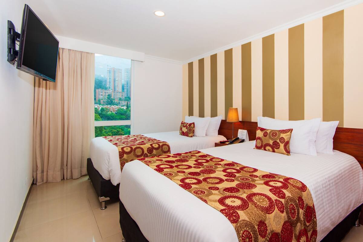 A hotel room with two beds, a TV, and a window with a view of buildings.