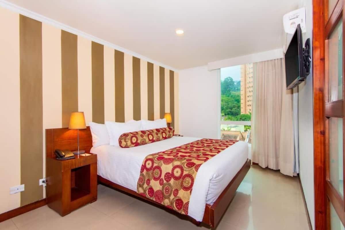 A hotel room with a large bed, a nightstand with a lamp, a TV mounted on the wall, and a balcony with curtains.