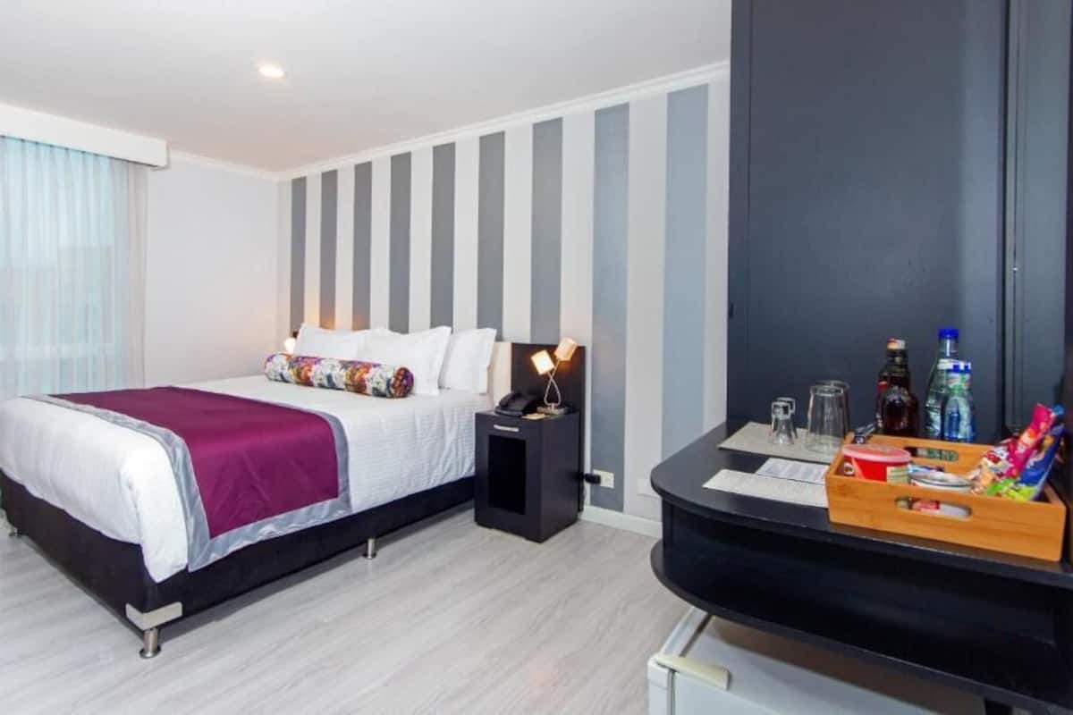 A hotel room with a bed, bedside table, and a small desk with a tray of snacks.
