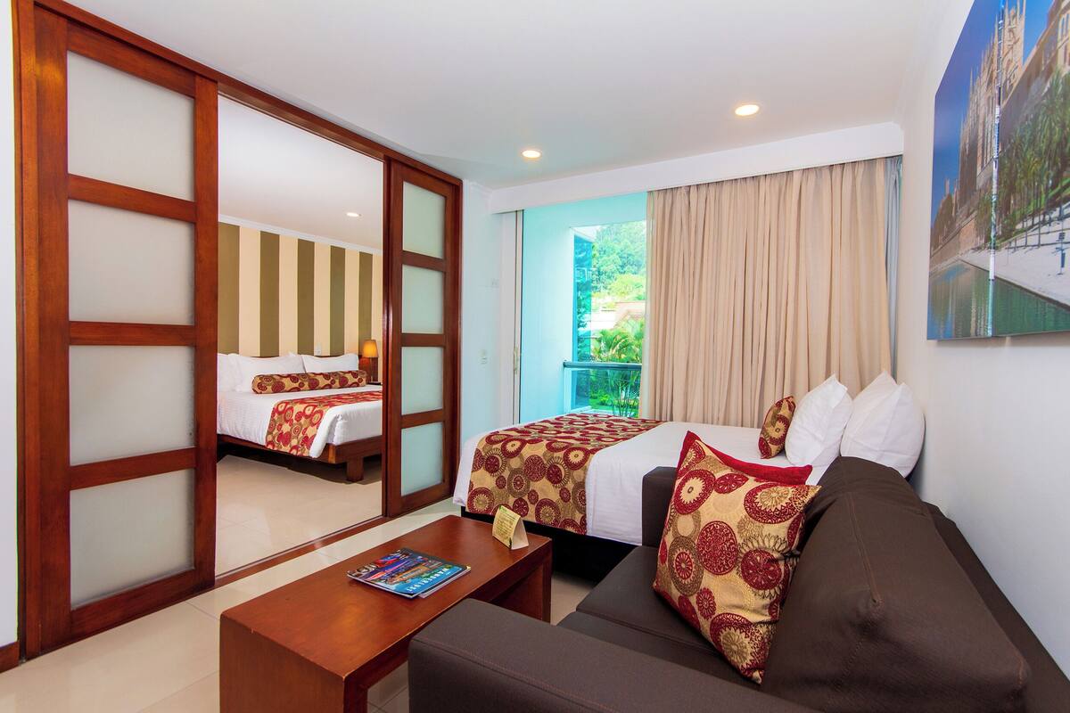 A hotel room with a bed, a sofa, a coffee table, a sliding door, and a view of greenery outside.