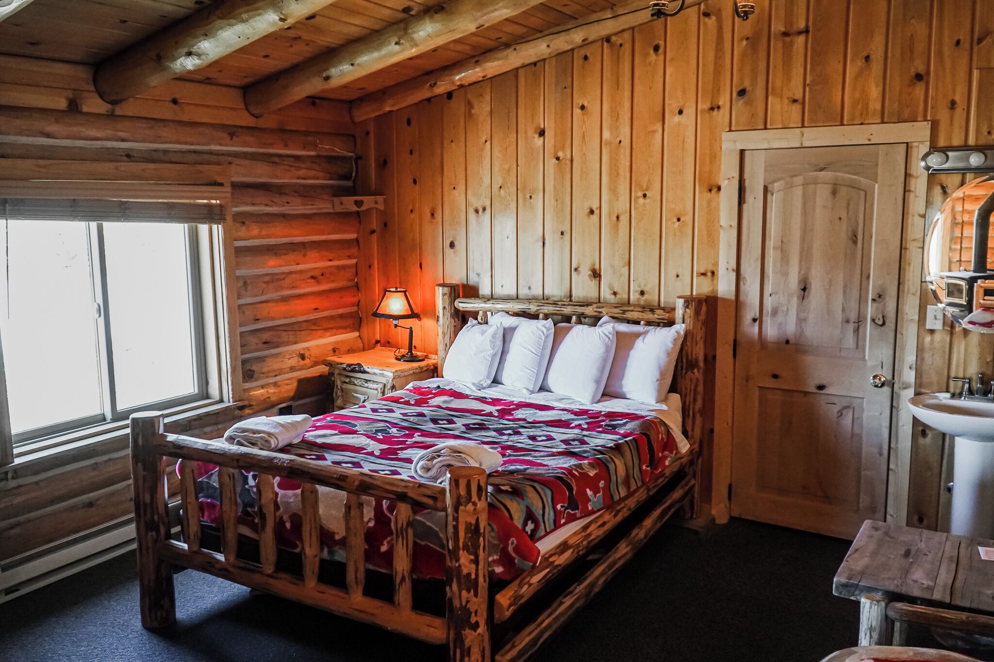 A wooden cabin bedroom with a bed, a nightstand, a lamp, a window, and a door.