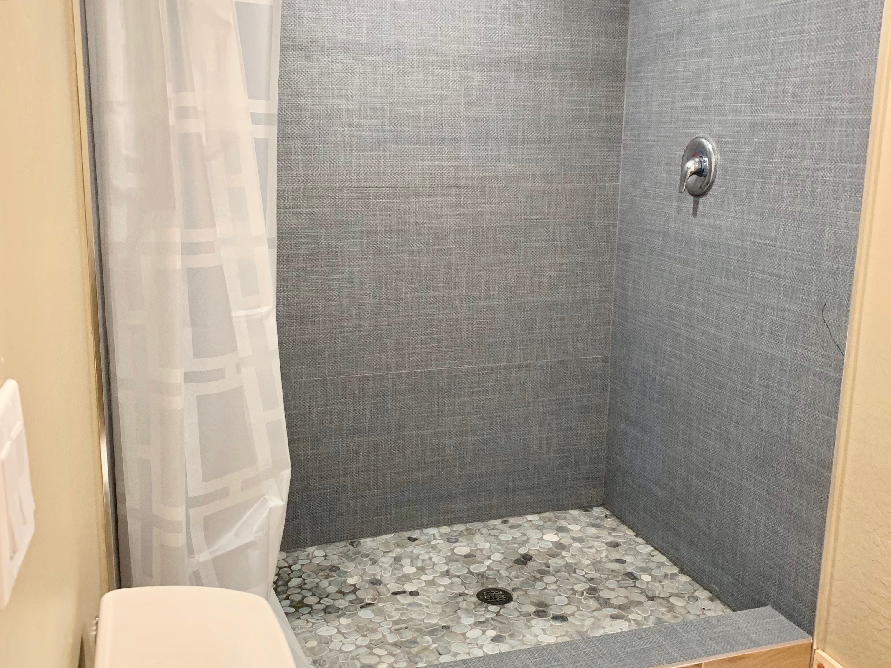 Un petit espace douche fermé, avec un mur gris texturé, un rideau de douche et un sol composé de petites pierres multicolores.
