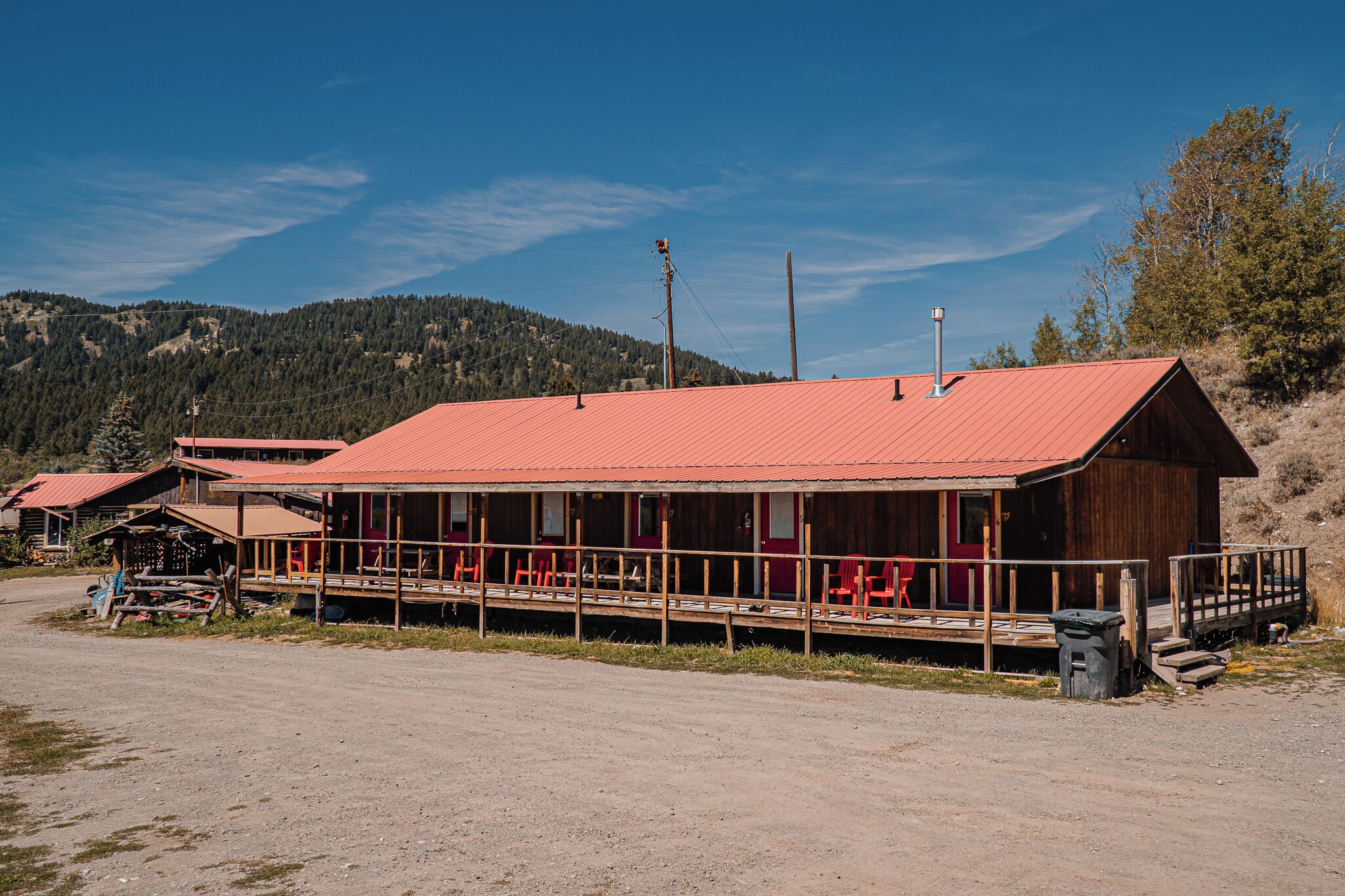 A wooden building with a red roof, a covered porch with red chairs, and a dirt ground.