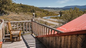 Una terraza de madera con una mecedora, un edificio de techo rojo y vistas a un río y montañas.