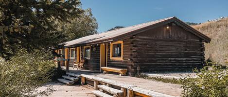 A rustic log cabin with a wooden deck and benches, surrounded by trees and a clear sky.