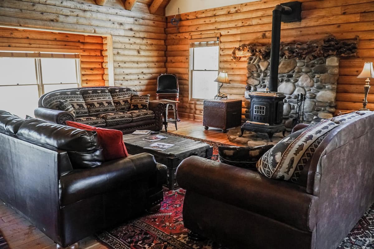 Interior de una cabaña de troncos con un sofá de cuero, sillones, una mesita, chimenea y una pared de piedra.