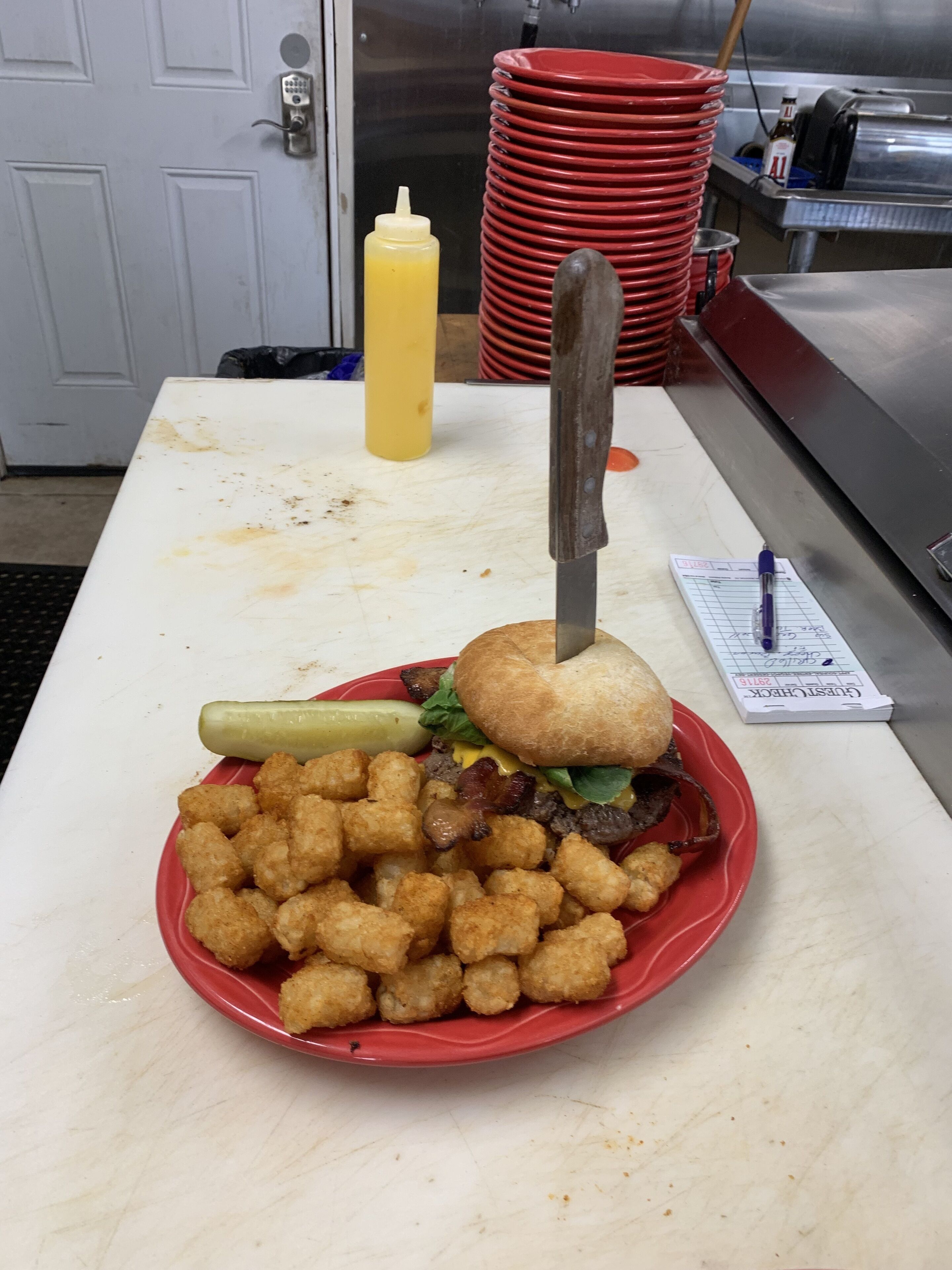 Un repas composé d’un hamburger, de pommes de terre en forme de petits cubes et d’un cornichon, le tout servi sur une assiette rouge posée sur une table.
