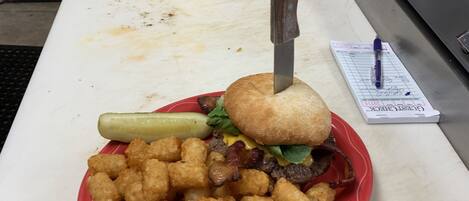 Un repas composé d’un hamburger, de pommes de terre en forme de petits cubes et d’un cornichon, le tout servi sur une assiette rouge posée sur une table.