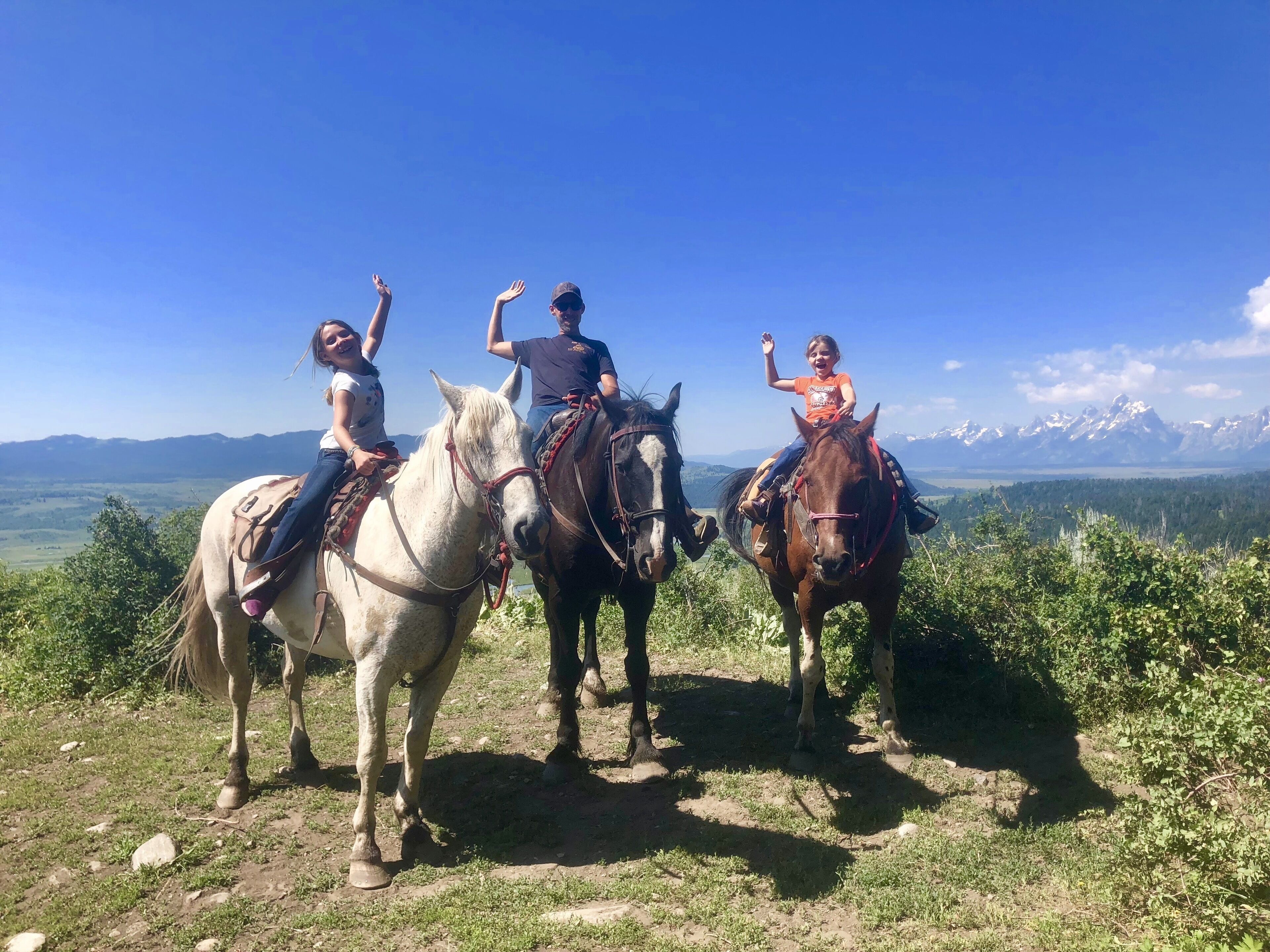 Three people on horseback on a grassy hill with a mountain range in the background.