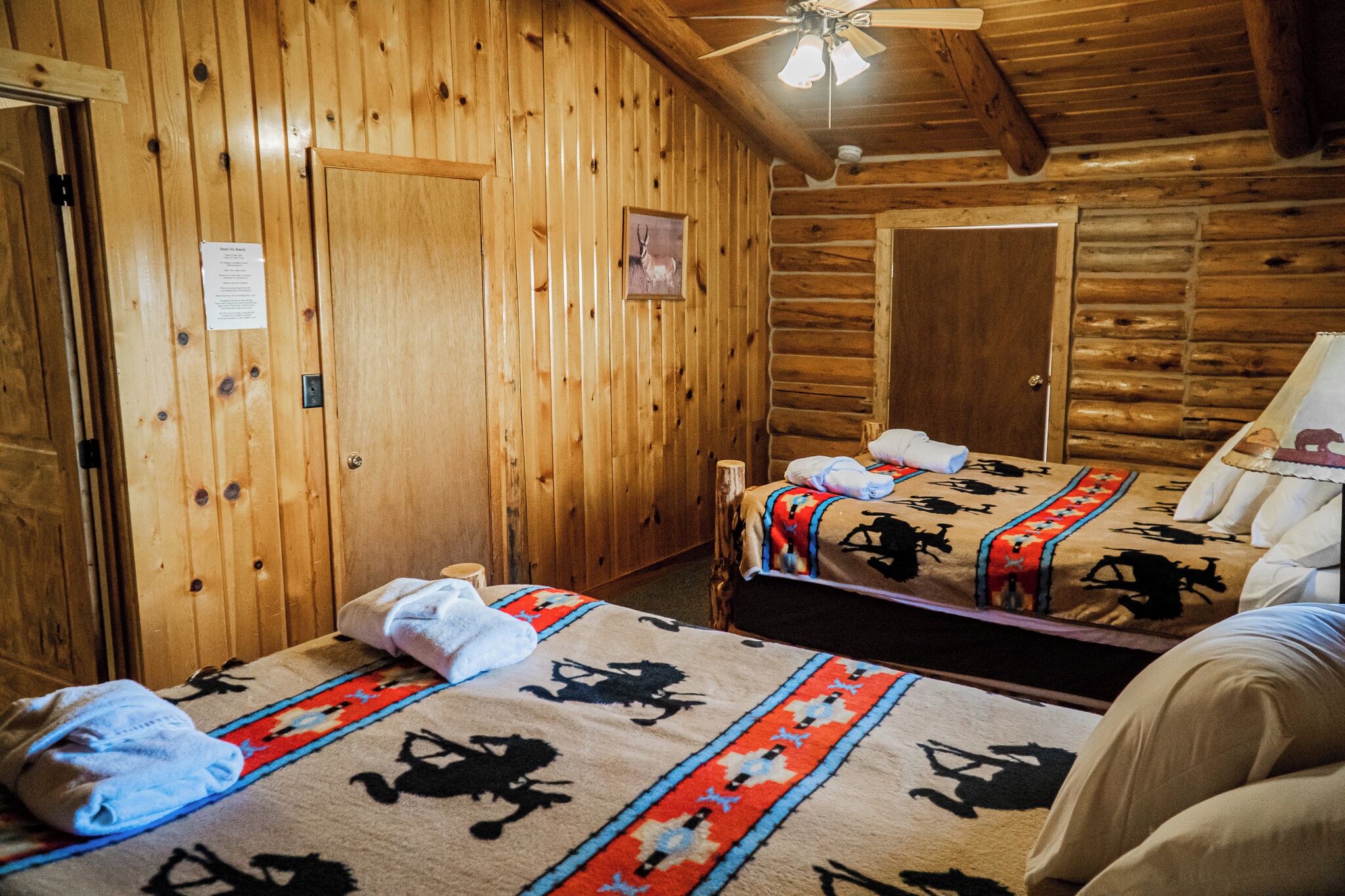 Quarto de cabana de madeira com duas camas, um ventilador de teto e uma porta.