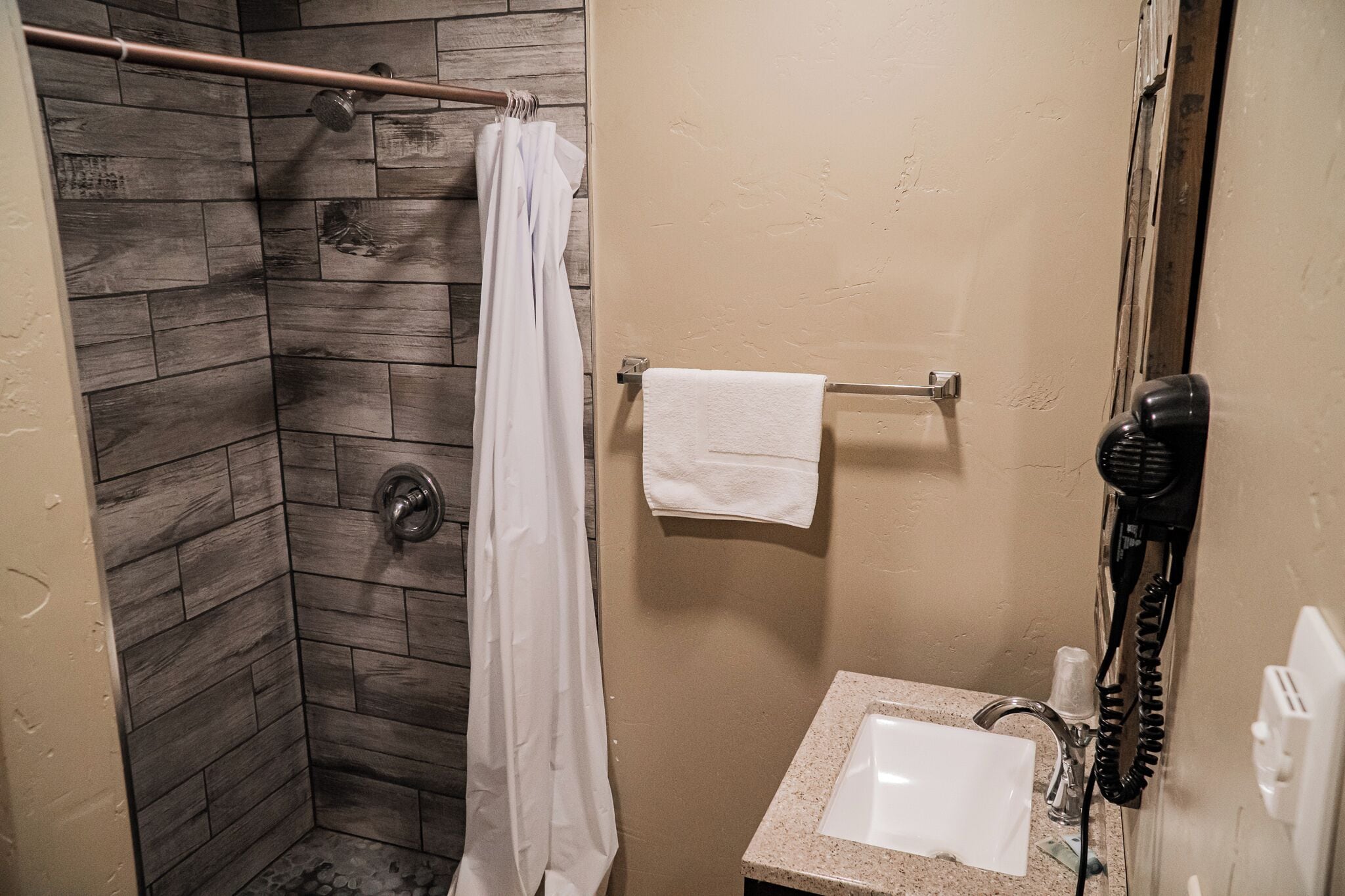 A bathroom with a shower area featuring wooden tiles, a white towel hanging on a rack, a hairdryer, and a sink with a mirror.