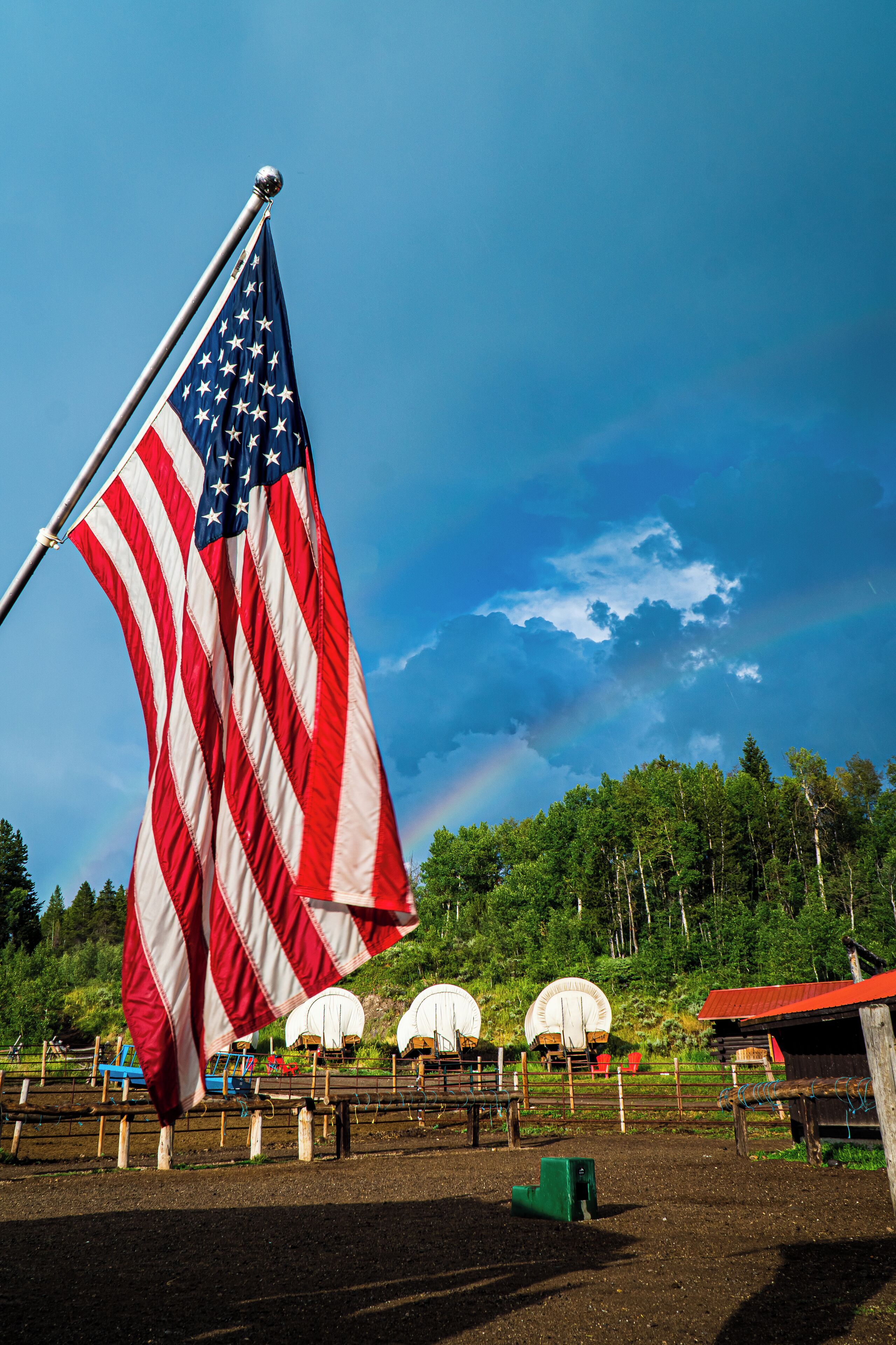 A flagpole with an American flag, a dirt ground, and a fence with white tents in the background.