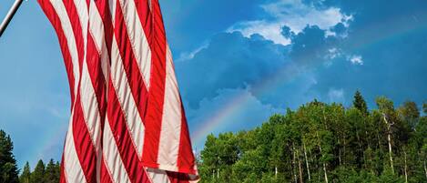 A flagpole with an American flag, a dirt ground, and a fence with white tents in the background.