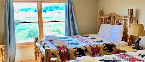 A room with two beds, a wooden headboard, a window with curtains, a lamp, and a view of greenery outside.