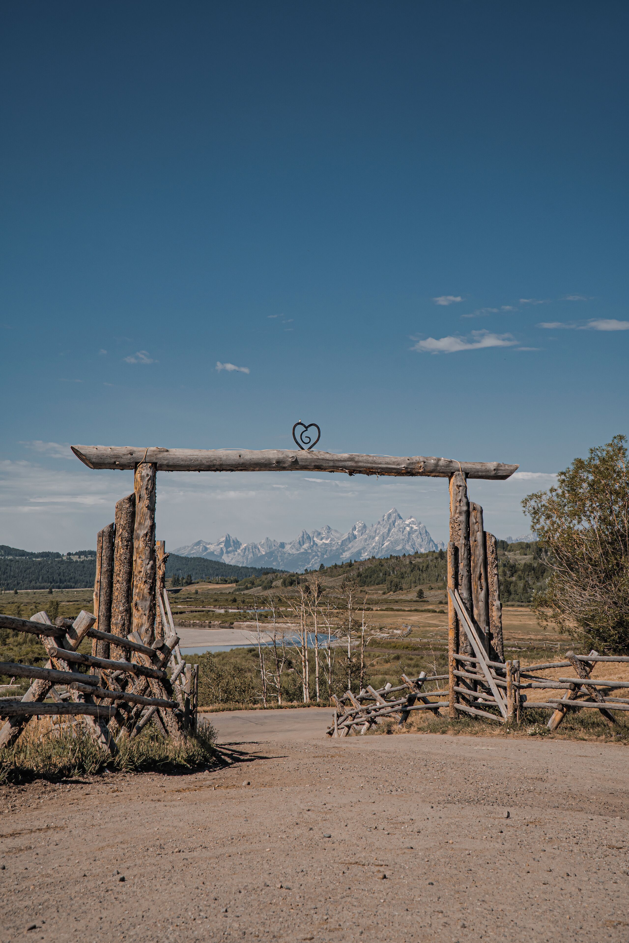 A rustic wooden gate with a heart symbol, leading to a mountainous landscape.