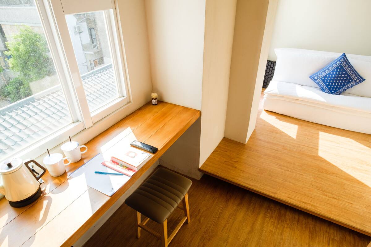 A small, bright room with a wooden desk, a white couch, and a window overlooking a cityscape.