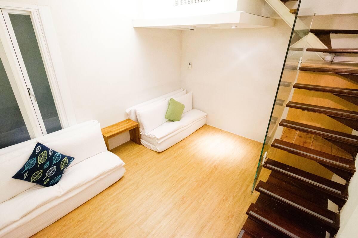 A compact living space with a wooden floor, a white sofa with a patterned pillow, a small bedside table, and a glass staircase.