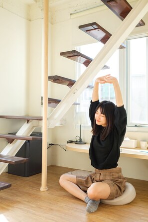 A person sitting on a cushion in a room with a wooden floor, a desk, and a staircase with wooden steps.