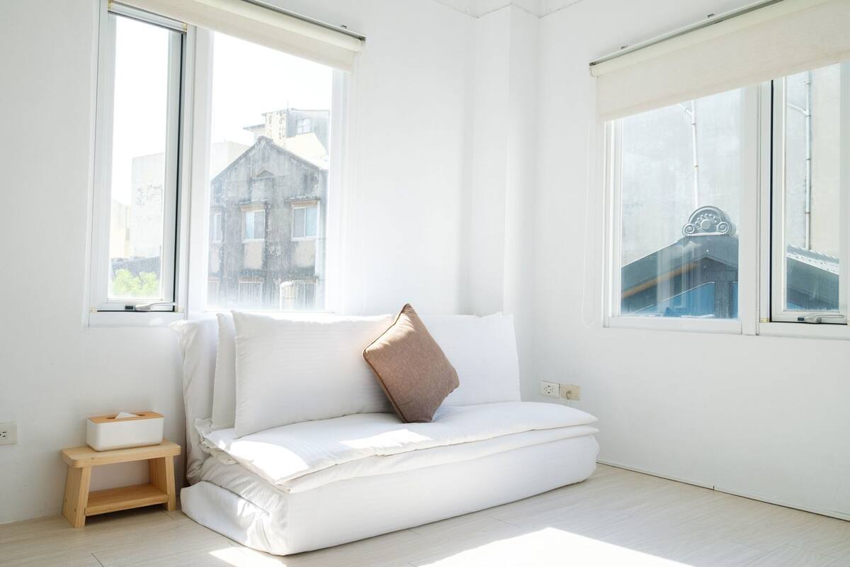 A bright room with a white sofa, a small wooden stool, and two windows with views of buildings.