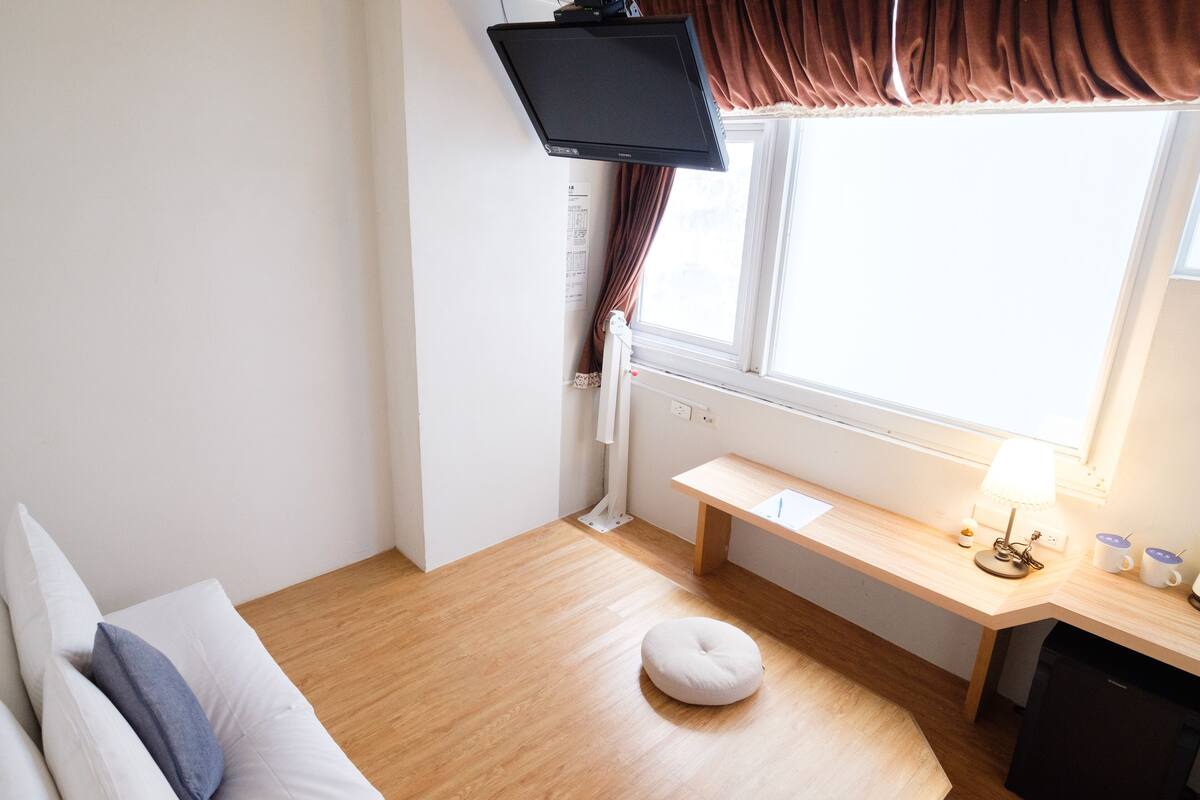 A room with a wooden floor, a white sofa with cushions, a small round ottoman, a wooden desk with a lamp, and a television mounted on the wall.