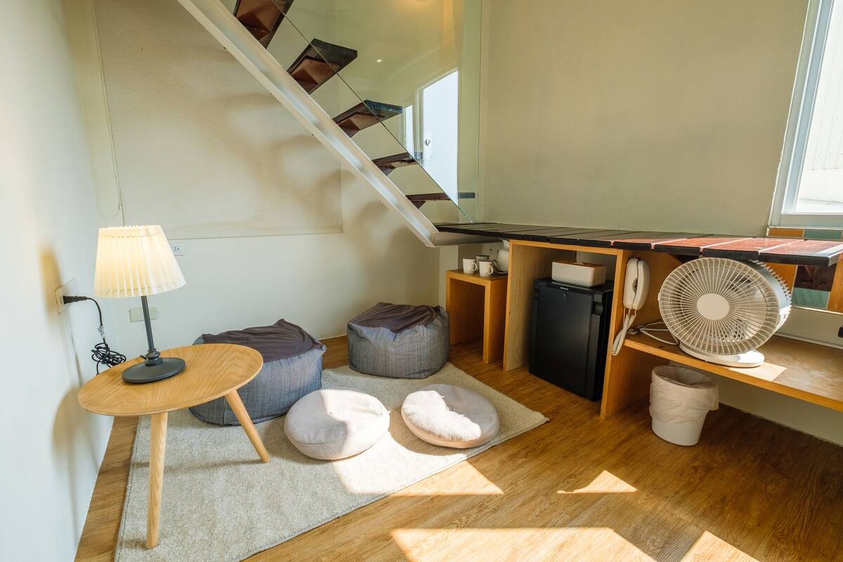 A compact living space with a staircase, a small table with a lamp, bean bags, a fan, and a desk with a telephone.
