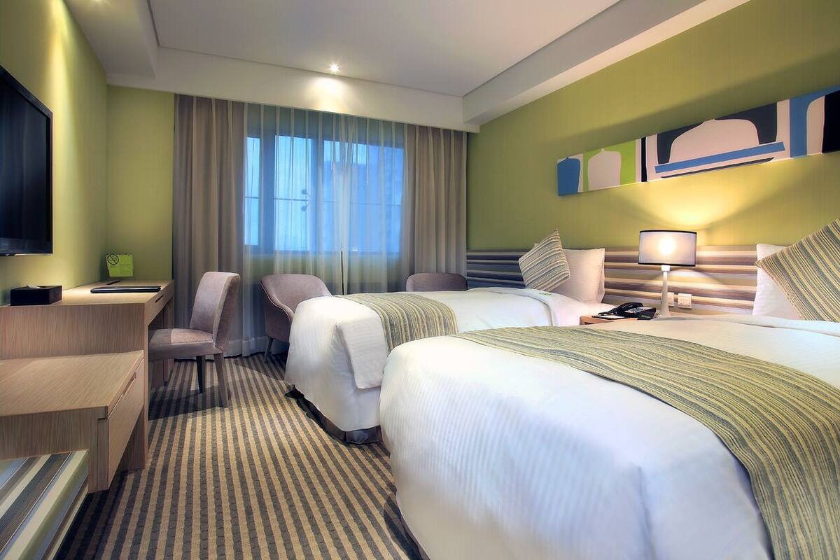 A hotel room with two beds, a desk, a chair, a TV, and a window with curtains.