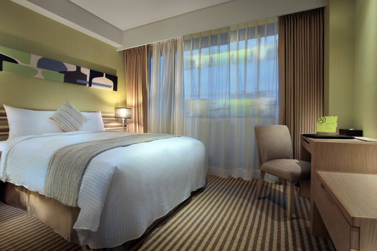 A hotel room with a large bed, a desk, a chair, and a view of the outdoors through sheer curtains.