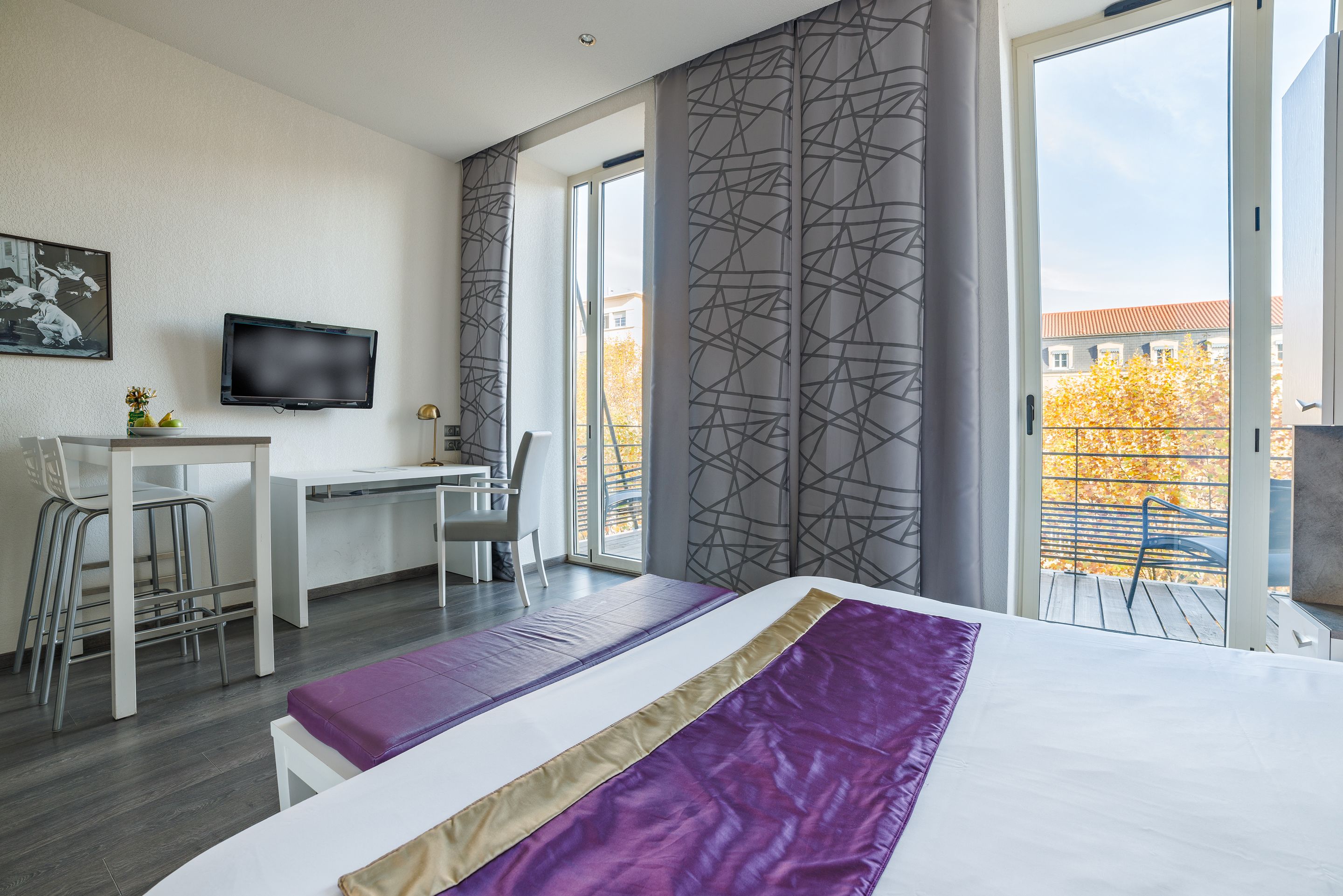 A modern hotel room with a bed, a desk, a chair, a TV, and a balcony with a view of buildings and trees.